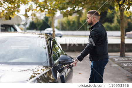 Handsome bearded guy opens a door of his luxury car on parking. Man finish work and drives home  119653336
