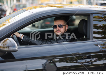 Success in motion. Handsome young man in leather jacket and sunglasses smiling while driving a car. Side view from outside  119653347