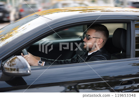 Success in motion. Handsome young man in leather jacket and sunglasses driving a car. Side view from outside  119653348