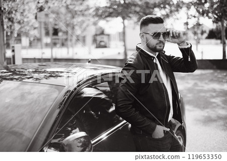 Portrait of a businessman standing beside his luxury car, placing his left hand on his sunglasses 119653350