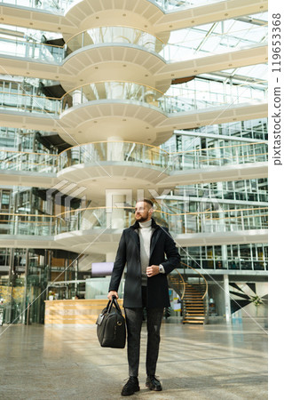 Man in coat walking though the big office. Guy hold travel bag. Stylish guy after working day Man in coat walking though the big office. Guy hold travel bag. Stylish guy after working day 119653368