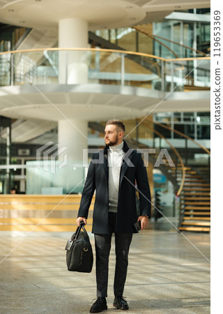 Man in coat walking though the big office. Guy hold travel bag. Stylish guy after working day Man in coat walking though the big office. Guy hold travel bag. Stylish guy after working day 119653369