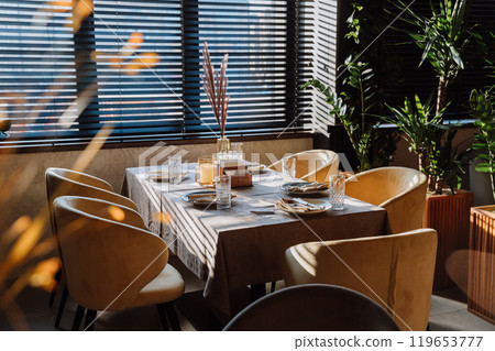 Elegant restaurant table setup with natural sunlight, cozy chairs, and stylish decor for dining Elegant restaurant table setup with natural sunlight, cozy chairs, and stylish decor for dining 119653777