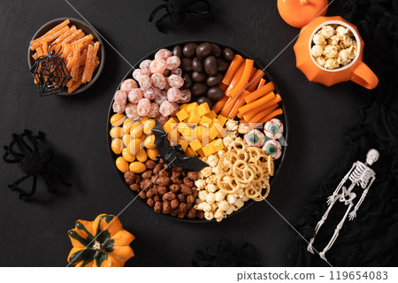 Halloween sweet charcuterie board with pumpkin, popcorn, orange chocolate candy, spider, crackers on black background. Scary decoration. Festive food for kids party. View from above. Halloween sweet charcuterie board with pumpkin, popcorn, orange chocolate candy, spider, crackers on black background. Scary decoration. Festive food for kids party. View from above. 119654083