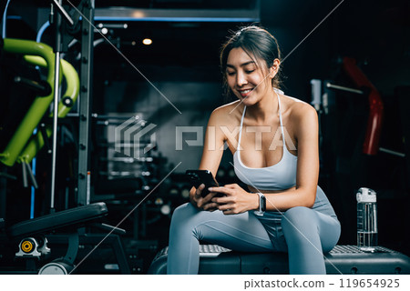 Asian woman resting on bench and checking all updates after exercising in gym interior, female using mobile phone application during training at fitness gym, sport workout healthy lifestyle concept 119654925