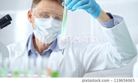 Scientist with protective gloves and glasses examining lab tube with a green fluid. Microbiology and science concept Scientist with protective gloves and glasses examining lab tube with a green fluid. Microbiology and science concept 119656065
