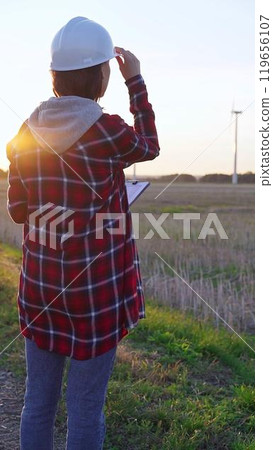 Woman engineer wearing a white protective helmet is taking notes with a clipboard in a field with wind turbines, as the sun sets. Clean energy and engineering concept 119656107