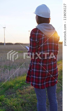Woman engineer wearing a white protective helmet is taking notes with a clipboard in a field with wind turbines, as the sun sets. Clean energy and engineering concept Woman engineer wearing a white protective helmet is taking notes with a clipboard in a field with wind turbines, as the sun sets. Clean energy and engineering concept 119656127