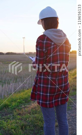 Woman engineer wearing a white protective helmet is taking notes with a clipboard in a field with wind turbines, as the sun sets. Clean energy and engineering concept Woman engineer wearing a white protective helmet is taking notes with a clipboard in a field with wind turbines, as the sun sets. Clean energy and engineering concept 119656133