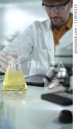 Man scientist wearing white protective gloves, and glasses is holding a yellow chemical solution inside an Erlenmeyer flask in a laboratory, vertical portrait. Science and medicine 119656213