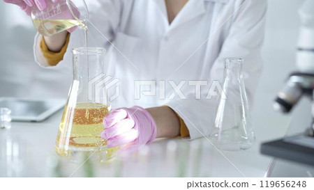 A scientist, wearing a lab coat and pink protective gloves, is pouring a yellow oily liquid from one beaker to another near microscope in laboratory, close up. Medicine and science 119656248