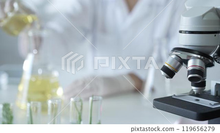 A scientist, wearing a lab coat and white protective gloves, is pouring a yellow oily liquid from one beaker to another near microscope in laboratory, close up. Medicine and science concept 119656279