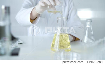 A scientist, wearing a lab coat and white protective gloves, is pouring a yellow oily liquid from one beaker to another in laboratory, close up. Medicine and science concept 119656289
