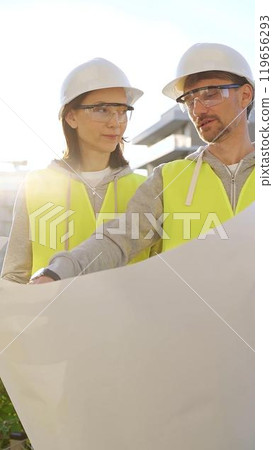 Two architects wearing safety hard hats and vests holding blueprint and discussing something on construction site at sunrise, front vertical view. Architecture engineering and teamwork concept 119656293