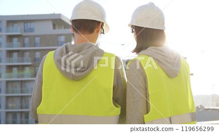 Two architects wearing safety hard hats and vests holding blueprint and discussing construction site project at sunrise, back view. Architecture, engineering and teamwork concept Two architects wearing safety hard hats and vests holding blueprint and discussing construction site project at sunrise, back view. Architecture, engineering and teamwork concept 119656299