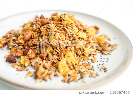 A bowl of granola with nuts and seeds on a white plate. The granola is a mix of different types of grains and nuts, and it looks like a healthy snack. 119667465