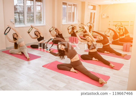 A group of six athletic women doing pilates or yoga on pink mats in front of a window in a beige loft studio interior. Teamwork, good mood and healthy lifestyle concept. A group of six athletic women doing pilates or yoga on pink mats in front of a window in a beige loft studio interior. Teamwork, good mood and healthy lifestyle concept. 119667466