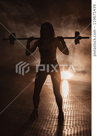 A powerful silhouette of a determined female weightlifter in a dynamic gym symbolizes athleticism, courage, and perseverance, embodying the spirit of a warrior pushing boundaries and defying limits 119671439