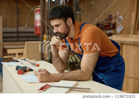 Pensive man carpenter reading blueprints and thinking during woodworking process Pensive man carpenter reading blueprints and thinking during woodworking process 119672831