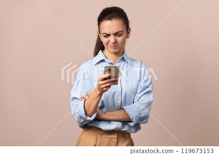 skeptical woman in blue shirt holding mobile phone looking at smartphone on beige background. skeptical woman in blue shirt holding mobile phone looking at smartphone on beige background. 119673153