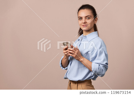 positive pretty woman using mobile phone looking at camera on beige background. copy space 119673154