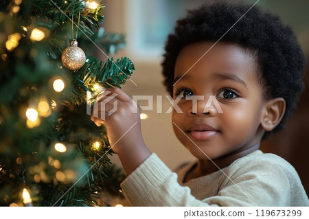 Adorable little African American boy with curly hair decorating Christmas tree with golden balls in living room at home, child toddler preparing to celebrate new year 119673299