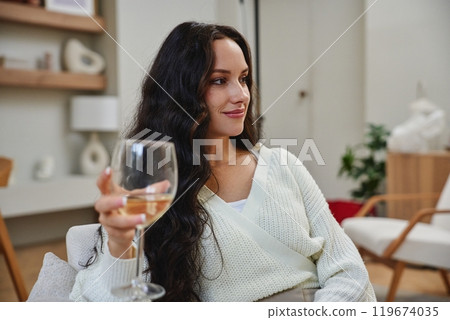 Woman relaxing indoors while holding a glass of white wine in a cozy setting Woman relaxing indoors while holding a glass of white wine in a cozy setting 119674035