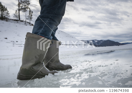 Close-up of person wearing waterproof boots walking on a frozen lake in a snowy landscape during winter. Thin ice, the danger of going out on the ice in winter 119674398