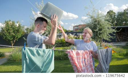 Middle-aged woman playfully placing a laundry basket on a young man's head as they hang clothes on a sunny day in a garden, enjoying a lighthearted moment together. 119675642