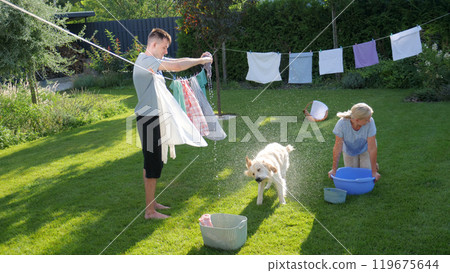 A man holds laundry while a Golden Retriever shakes off water, with a woman kneeling nearby, enjoying a sunny day in the backyard. 119675644