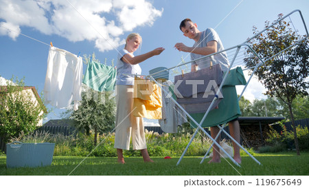 A woman and a man hanging clothes on a drying rack and clothesline in a bright backyard, enjoying a sunny day doing household chores together. A woman and a man hanging clothes on a drying rack and clothesline in a bright backyard, enjoying a sunny day doing household chores together. 119675649