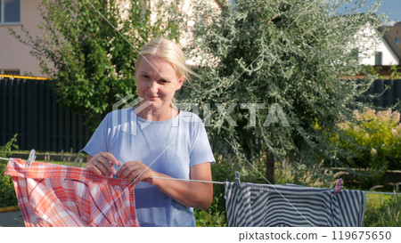A woman hanging a plaid shirt on a clothesline in a sunny backyard, surrounded by greenery. A woman hanging a plaid shirt on a clothesline in a sunny backyard, surrounded by greenery. 119675650
