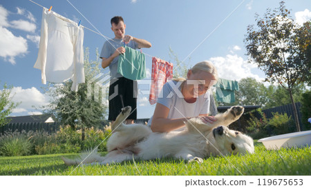 Woman petting a Golden Retriever on the grass while a man hangs clothes on a line in a sunny garden. Woman petting a Golden Retriever on the grass while a man hangs clothes on a line in a sunny garden. 119675653