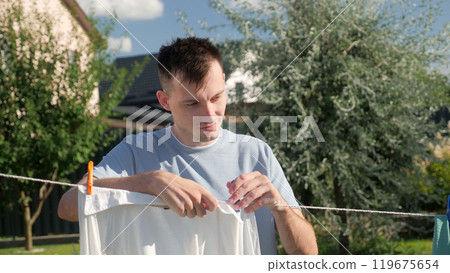 A young man pins a white shirt on a clothesline in a sunlit backyard, concentrating on the task. 119675654