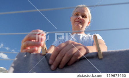 A woman secures clothes on a line with clothespins, viewed from a low angle against a clear blue sky. 119675660