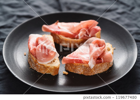 ciabatta with thin prosciutto on black plate on linen background 119677480
