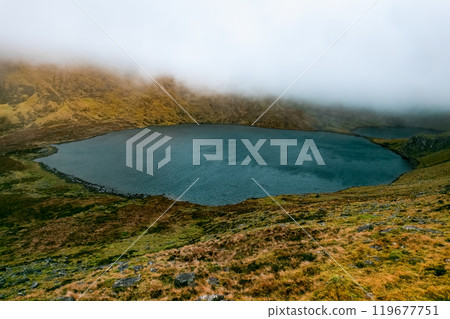 The tranquil lake reflects the surrounding mountains, partially obscured by clouds. Lush greenery and rocky terrain create a peaceful natural setting during dawn. 119677751