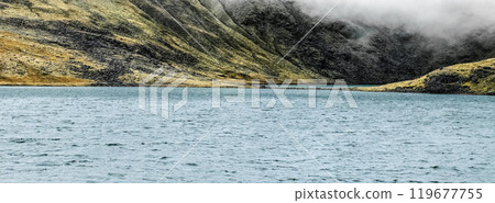 Calm waters reflect the rugged mountains and overcast skies in a tranquil setting, highlighting the untouched beauty of this remote landscape. 119677755