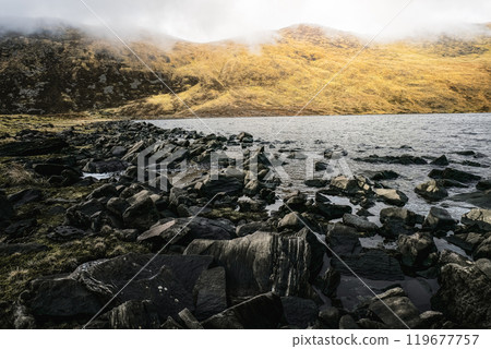 A rocky shoreline lies beside a serene lake, with grassy slopes rising in the background under a cloudy sky. The tranquil atmosphere invites reflection. A rocky shoreline lies beside a serene lake, with grassy slopes rising in the background under a cloudy sky. The tranquil atmosphere invites reflection. 119677757