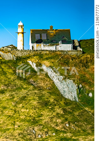 A white lighthouse stands on a rocky cliff. The sky is a clear blue, and the sun is shining brightly. A white lighthouse stands on a rocky cliff. The sky is a clear blue, and the sun is shining brightly. 119677772