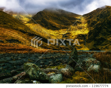 A small, still lake is nestled between two mountain slopes, covered in wispy clouds and a scattering of large, rounded rocks. A small, still lake is nestled between two mountain slopes, covered in wispy clouds and a scattering of large, rounded rocks. 119677774