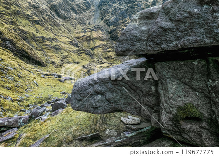 A unique rock formation shaped like a creature juts out from a hillside covered in vibrant green grass, surrounded by steep, rocky terrain and clouds above. A unique rock formation shaped like a creature juts out from a hillside covered in vibrant green grass, surrounded by steep, rocky terrain and clouds above. 119677775