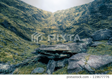 The lush valley is surrounded by steep rocky cliffs, with mist hanging low, creating an atmospheric and tranquil setting with scattered boulders. 119677776