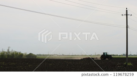 Outside the city a tractor plows a field with a special tool against a background of trees and power lines. Outside the city a tractor plows a field with a special tool against a background of trees and power lines. 119678259