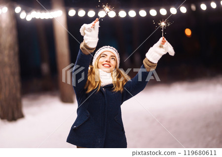Cheerful young woman holding a sparkler in hand in forest. Woman happiness and playing firework. Cheerful young woman holding a sparkler in hand in forest. Woman happiness and playing firework. 119680611