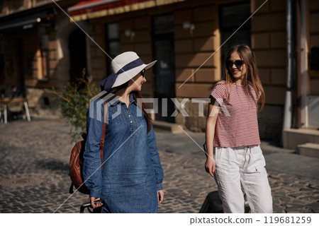 carrying suitcase, city travel, sunny urban. On bright day in city, two young women in casual attire walk along cobblestone street with suitcases. carrying suitcase, city travel, sunny urban. On bright day in city, two young women in casual attire walk along cobblestone street with suitcases. 119681259
