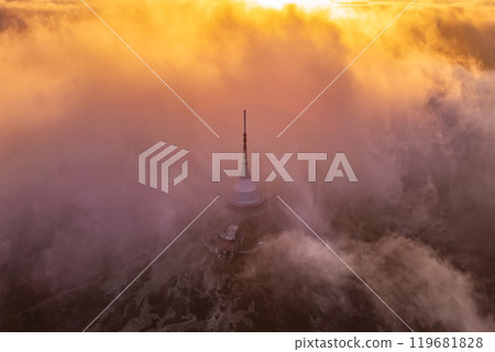 The sunset casts a warm, golden hue over Jested mountain in Liberec, Czechia, as clouds swirl around the tower, creating a mystical atmosphere during this serene evening. 119681828
