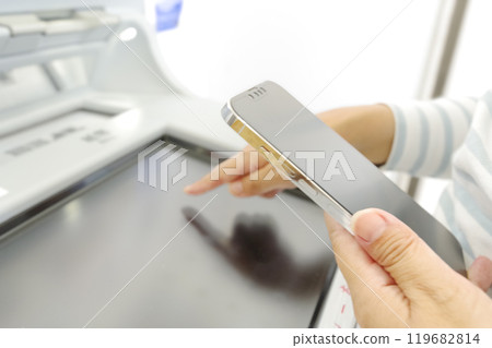 一位女士一邊看智慧型手機一邊操作 ATM 119682814