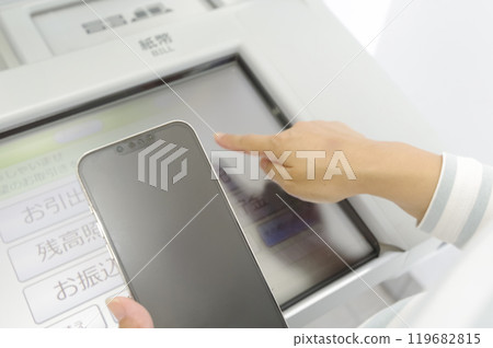 一位女士一邊看智慧型手機一邊操作 ATM 一位女士一邊看智慧型手機一邊操作 ATM 119682815