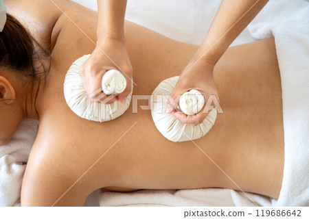 Close-up of woman getting back massage with two herbal compresses at spa bed, relaxation and traditional wellness techniques on white towel 119686642